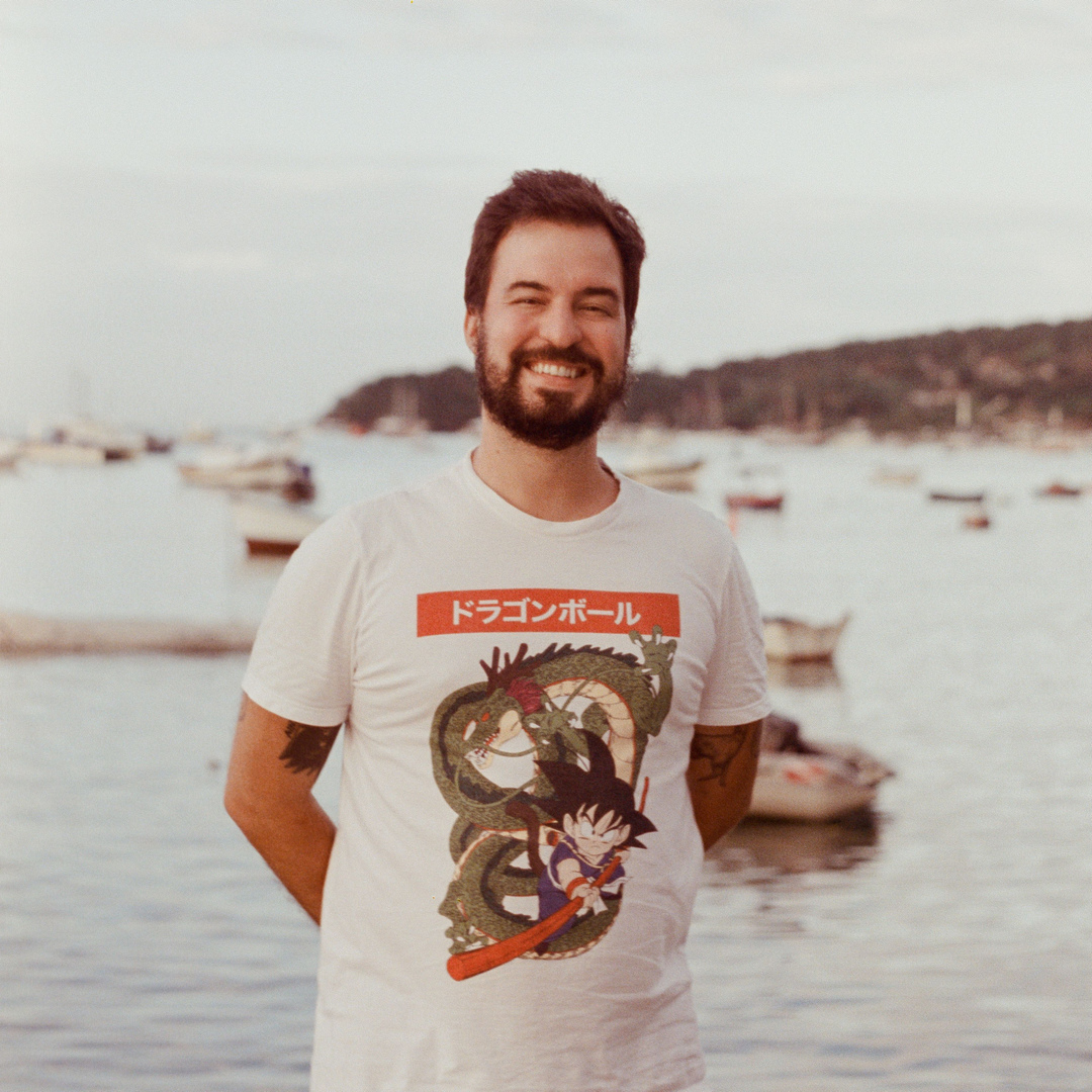 foto analógica do Nigel Goodman sorrindo, à frente de uma praia com barcos ancorados